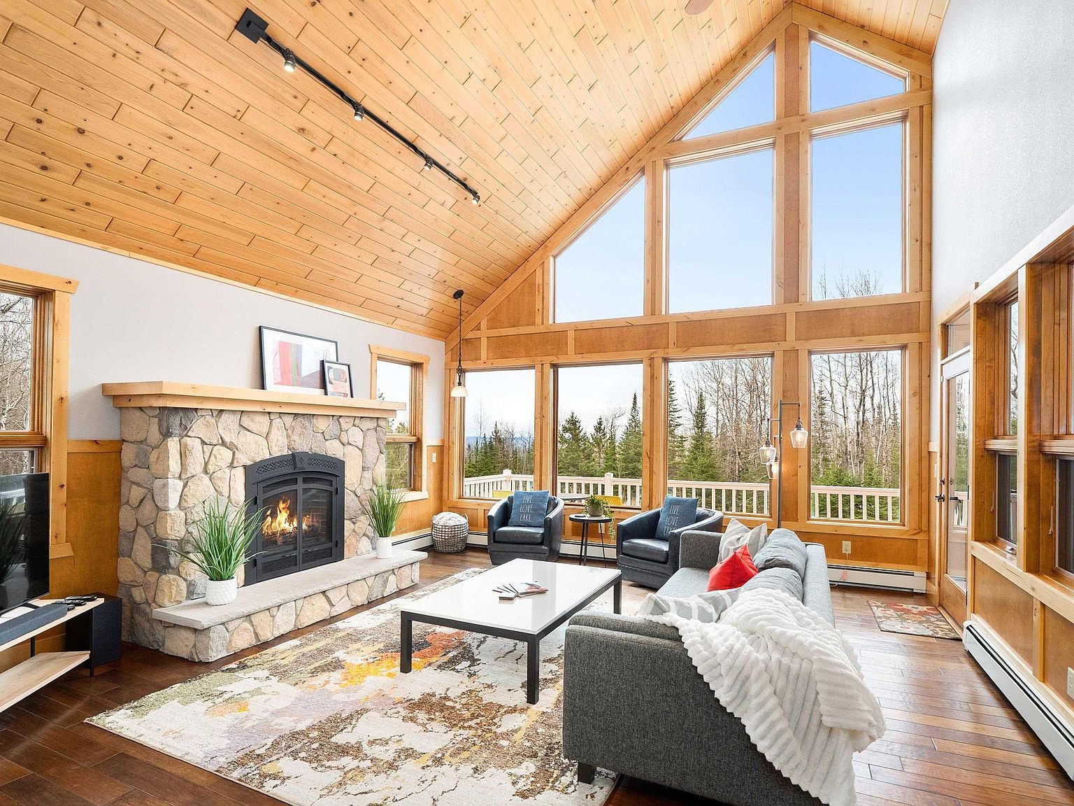 Great room with vaulted knotty pine ceiling, stone fireplace and a two-story wall of windows facing the forest